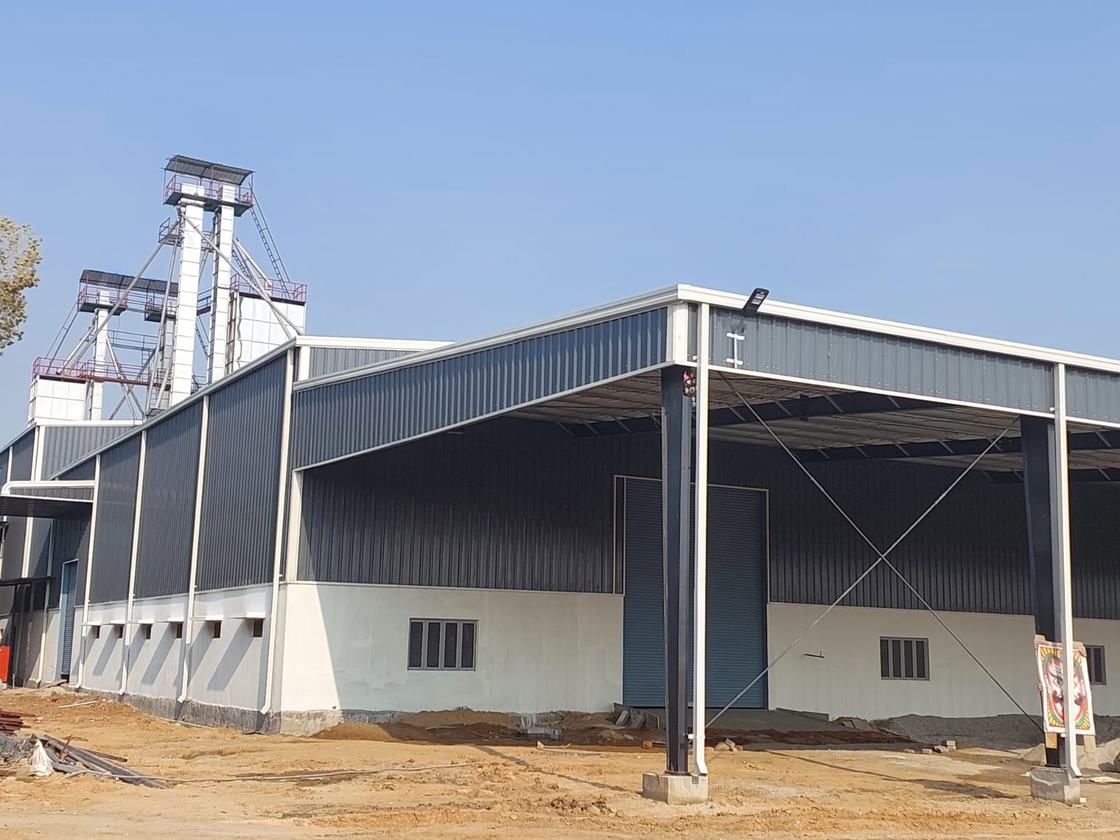 Steel-framed industrial shed under construction with grain elevator behind